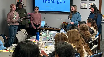 Group of Girl Scouts presenting in a meeting room while others watch and take notes.