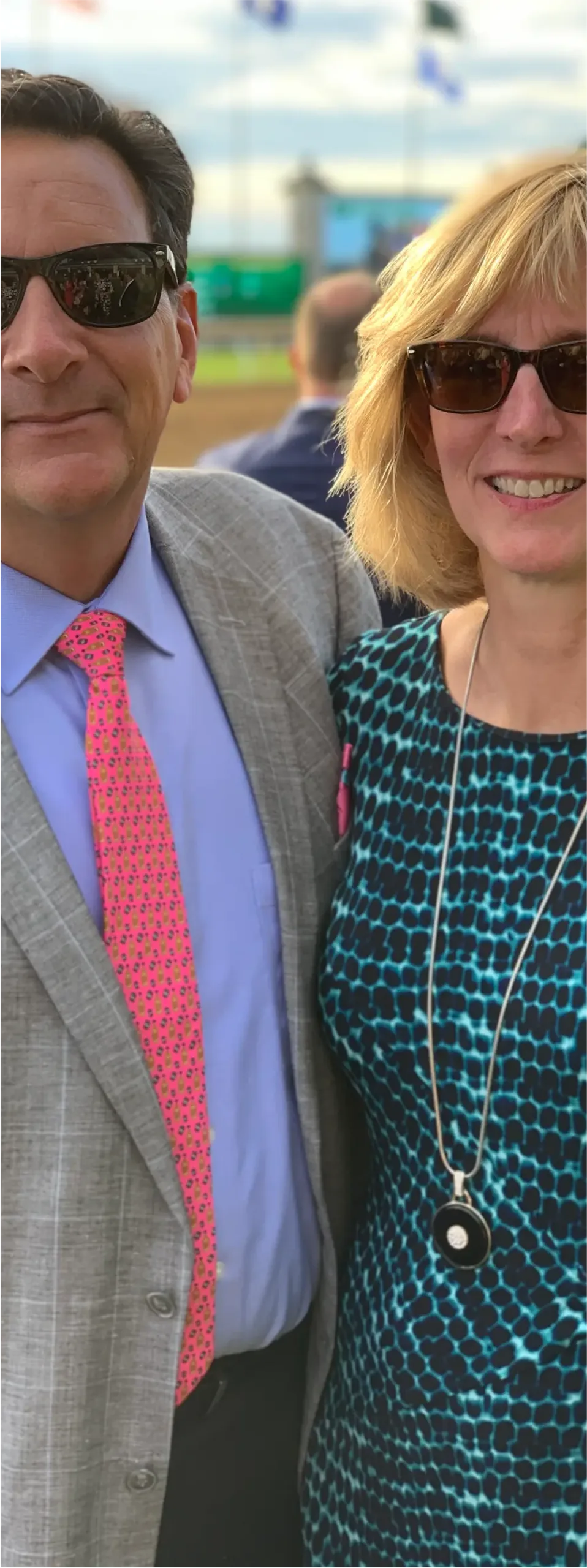 A man in a gray suit with a pink tie and a woman in a blue patterned dress posing outdoors together.