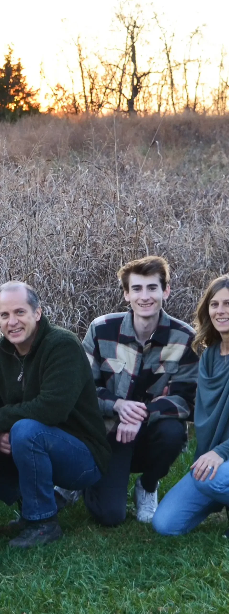 Three members of the Jackman family kneeling on grass with dry field and sunset trees in the background.