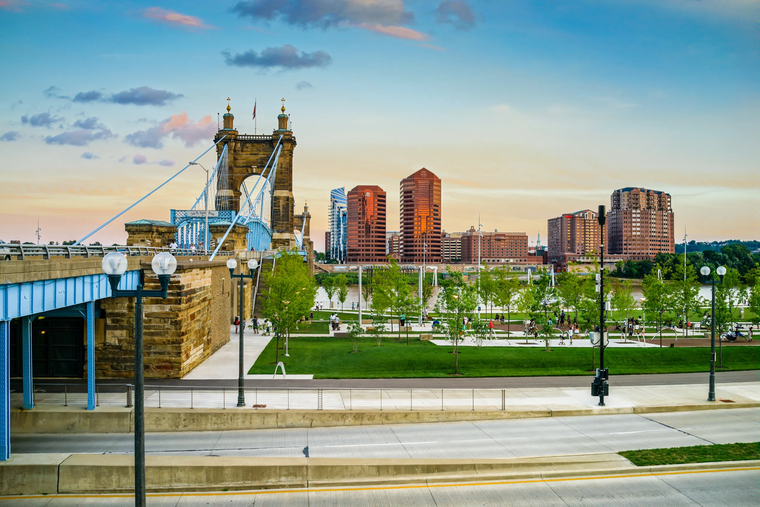 View of NKY and Roebling Suspension bridge leading into a cityscape with modern buildings at sunset.