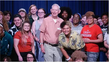 Roger Grein posing with a diverse group of smiling students on stage.