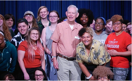 Roger Grein posing with a diverse group of smiling students on stage.